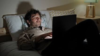 Young hispanic man using laptop lying on bed working at bedroom