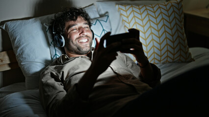 Fototapeta premium Young hispanic man watching video on smartphone lying on bed at bedroom