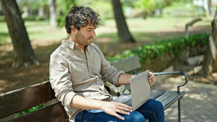 Obraz premium Young hispanic man using laptop sitting on bench at park