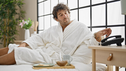 Young hispanic man having breakfast holding telephone at bedroom