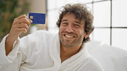 Young hispanic man wearing bathrobe holding credit card at bedroom