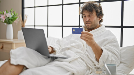 Young hispanic man wearing bathrobe shopping with laptop and credit card at bedroom - obrazy, fototapety, plakaty