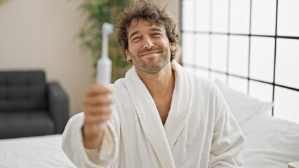Young hispanic man wearing bathrobe holding electric toothbrush smiling at bedroom