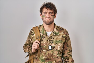 Hispanic young man wearing camouflage army uniform puffing cheeks with funny face. mouth inflated...