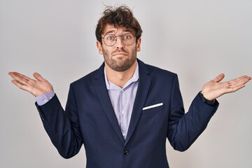 Hispanic business man wearing glasses clueless and confused expression with arms and hands raised. doubt concept.