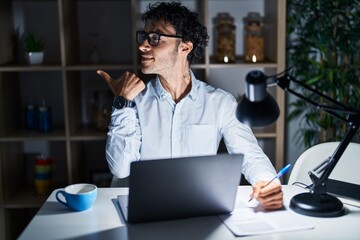 Hispanic man working at the office at night smiling with happy face looking and pointing to the side with thumb up.