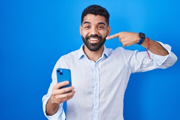 Hispanic man with beard using smartphone typing message smiling cheerful showing and pointing with fingers teeth and mouth. dental health concept.