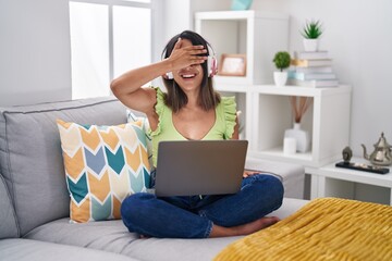 Hispanic young woman using laptop at home smiling and laughing with hand on face covering eyes for surprise. blind concept.