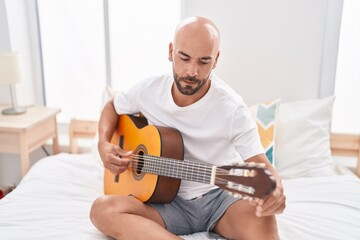 Young bald man playing classical guitar sitting on bed at bedroom