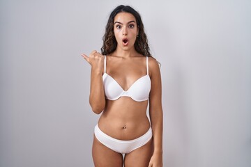 Young hispanic woman wearing white lingerie surprised pointing with hand finger to the side, open...