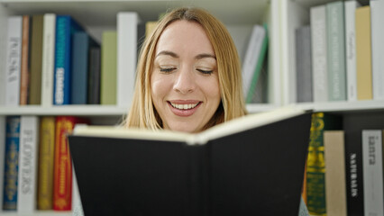 Young blonde woman student smiling confident reading book at library university