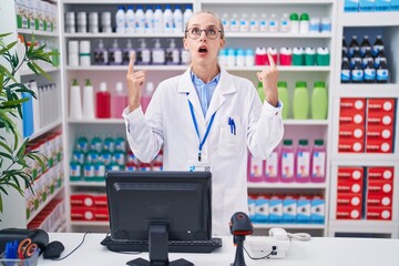 Young caucasian woman working at pharmacy drugstore amazed and surprised looking up and pointing with fingers and raised arms.