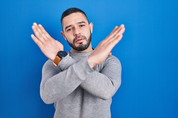 Hispanic man standing over blue background rejection expression crossing arms doing negative sign,...