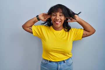 Hispanic woman standing over blue background smiling pulling ears with fingers, funny gesture....