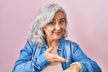Middle age woman with grey hair standing over pink background in hurry pointing to watch time,...