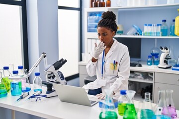 Young african american with braids working at scientist laboratory with laptop covering mouth with...