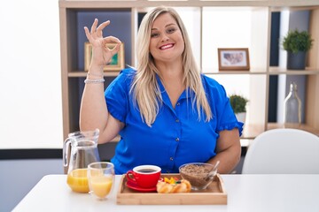 Caucasian plus size woman eating breakfast at home smiling positive doing ok sign with hand and fingers. successful expression.