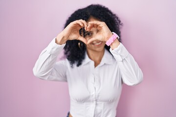 Hispanic woman with curly hair standing over pink background doing heart shape with hand and fingers smiling looking through sign