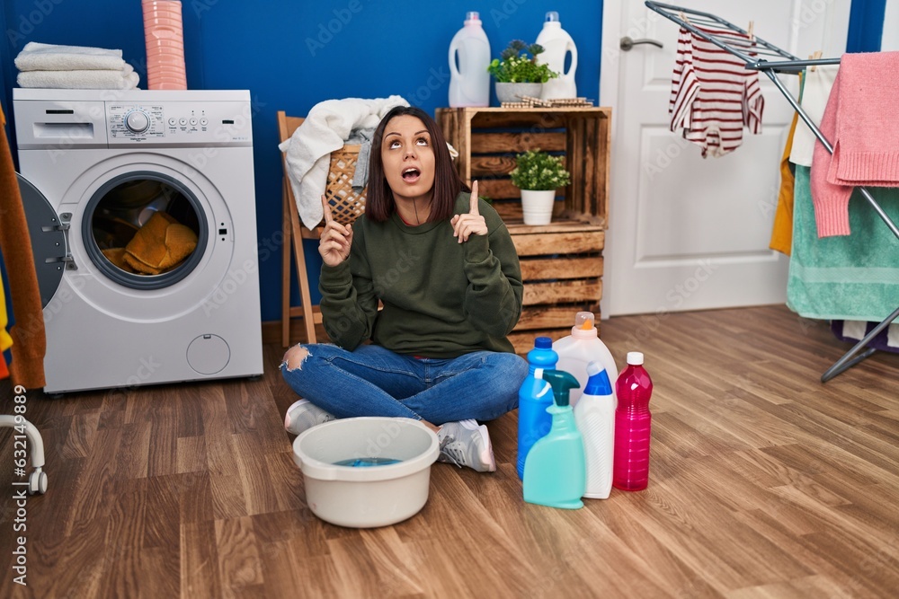 Sticker Young hispanic woman doing laundry washing by hand amazed and surprised looking up and pointing with fingers and raised arms.