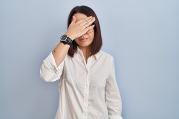 Young hispanic woman standing over white background covering eyes with hand, looking serious and...