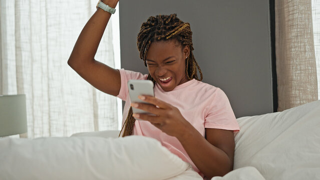 African american woman using smartphone sitting on bed celebrating at bedroom