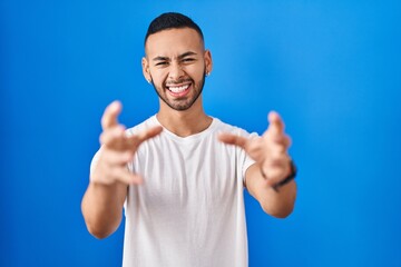 Fototapeta premium Young hispanic man standing over blue background shouting frustrated with rage, hands trying to strangle, yelling mad