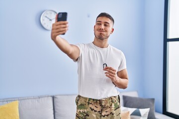 Young hispanic man wearing camouflage army uniform taking selfie at home puffing cheeks with funny...