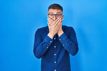 Young hispanic man wearing glasses over blue background shocked covering mouth with hands for mistake. secret concept.