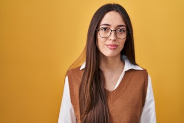Young brunette woman standing over yellow background wearing glasses relaxed with serious expression on face. simple and natural looking at the camera.