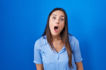 Young brunette woman standing over blue background in shock face, looking skeptical and sarcastic,...