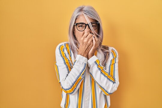 Middle Age Woman With Grey Hair Standing Over Yellow Background Wearing Glasses Smelling Something Stinky And Disgusting, Intolerable Smell, Holding Breath With Fingers On Nose. Bad Smell