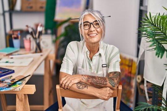 Middle age grey-haired woman artist smiling confident sitting with arms crossed gesture at art studio