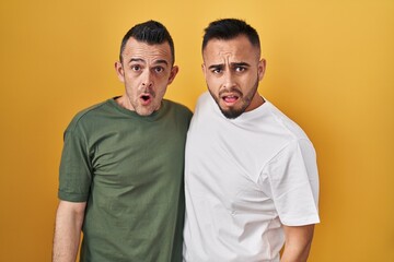 Homosexual couple standing over yellow background in shock face, looking skeptical and sarcastic, surprised with open mouth