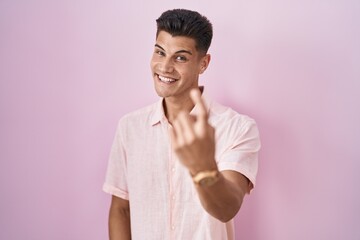 Young hispanic man standing over pink background beckoning come here gesture with hand inviting welcoming happy and smiling