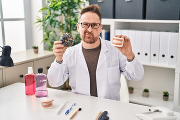 Middle age caucasian dentist man holding denture and doughnuts skeptic and nervous, frowning upset because of problem. negative person.