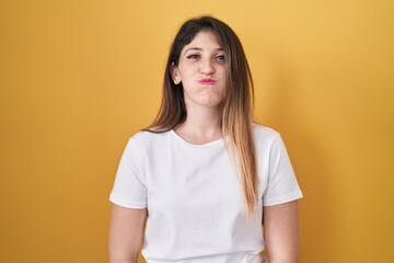 Young brunette woman standing over yellow background puffing cheeks with funny face. mouth inflated with air, crazy expression.