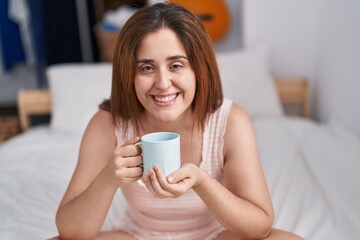 Young woman drinking cup of coffee sitting on bed at bedroom