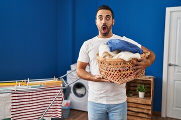 Young hispanic man with beard holding laundry basket scared and amazed with open mouth for...