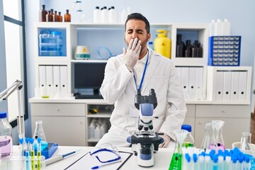 Young hispanic man with beard working at scientist laboratory bored yawning tired covering mouth with hand. restless and sleepiness.