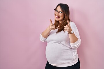 Pregnant woman standing over pink background pointing fingers to camera with happy and funny face. good energy and vibes.