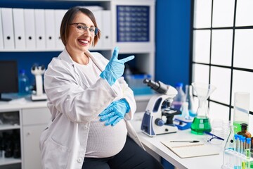 Pregnant woman working at scientist laboratory cheerful with a smile on face pointing with hand and finger up to the side with happy and natural expression