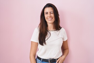 Young brunette woman standing over pink background winking looking at the camera with sexy...