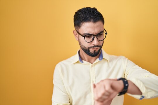 Hispanic young man wearing business clothes and glasses checking the time on wrist watch, relaxed and confident