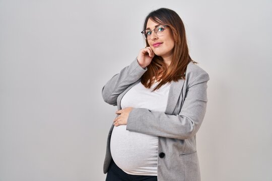 Pregnant business woman standing over white background serious face thinking about question with hand on chin, thoughtful about confusing idea