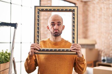 Young bald man with beard moving to a new home putting face inside vintage frame puffing cheeks with funny face. mouth inflated with air, catching air. - obrazy, fototapety, plakaty