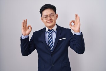 Young asian man wearing business suit and tie relaxed and smiling with eyes closed doing meditation gesture with fingers. yoga concept.