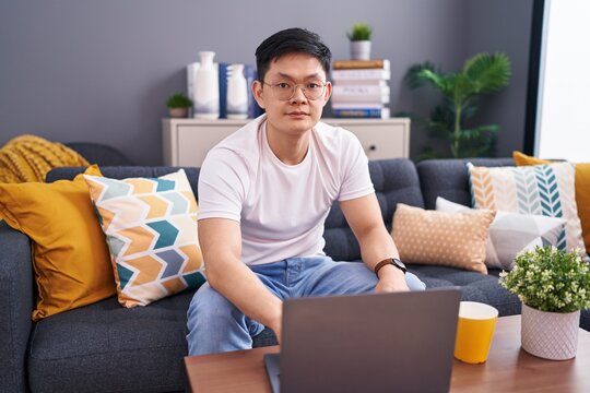 Young asian man using laptop at home sitting on the sofa relaxed with serious expression on face. simple and natural looking at the camera.