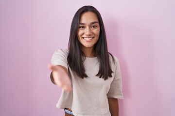 Young hispanic woman standing over pink background smiling cheerful offering palm hand giving assistance and acceptance.