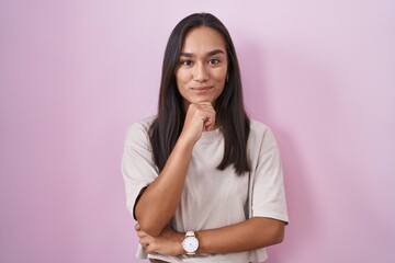 Young hispanic woman standing over pink background looking confident at the camera smiling with...