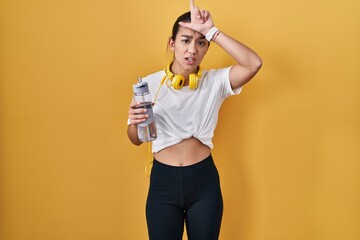 Young south asian woman wearing sportswear drinking water making fun of people with fingers on...